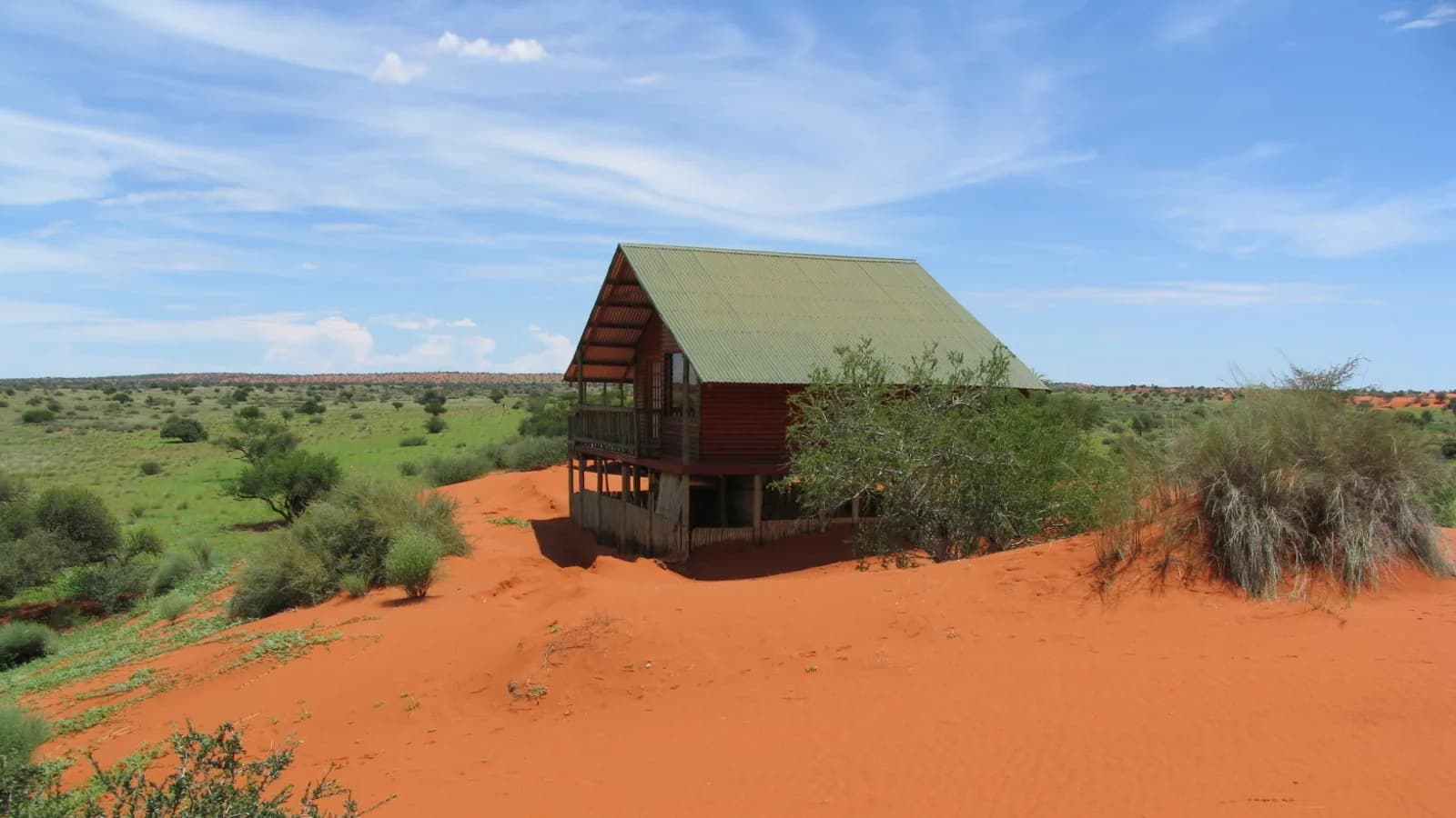 Houtlodge-kabin met 'n groen dak wat op rooi Kalahari-sand staan onder 'n helderblou hemel