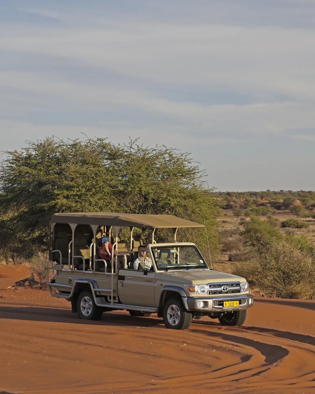 Ope safarivoertuig met twee gaste wat oor rooi sand ry tydens goue ure