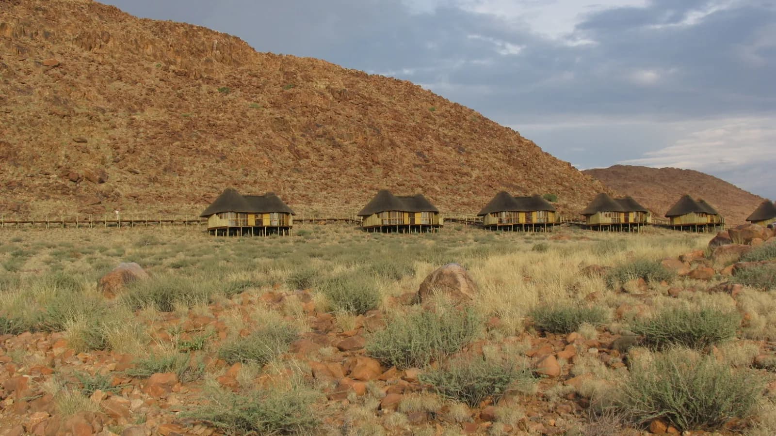Ry van rietdak chalets op palings onder ’n rooi rotshelling